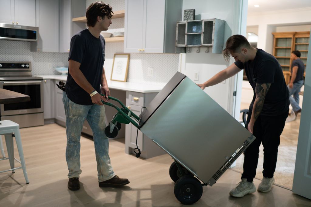 Two Loaders moving a stainless steel appliance on a hand truck through a kitchen