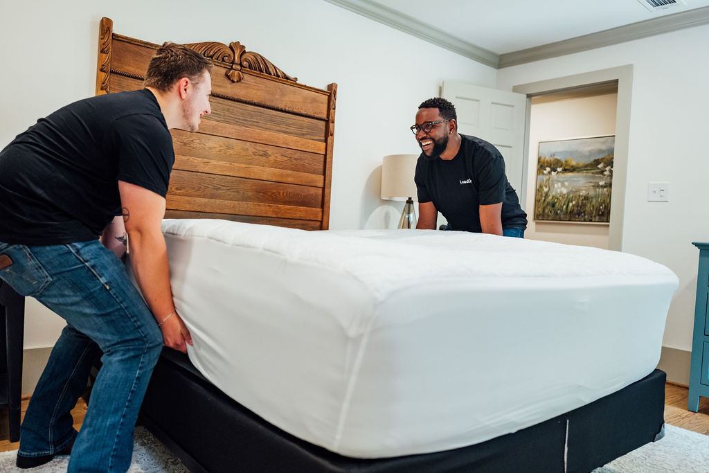 Two smiling Loaders lifting a mattress beside a wooden headboard