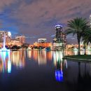 A panorama skyline photo of Orlando, Florida taken from across the water and the buildings reflecting in bright white, orange and yellow on the water.